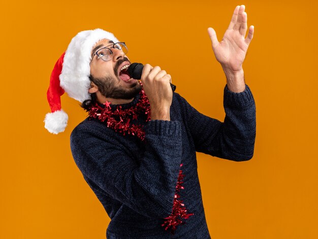 Bang opzoeken jonge knappe kerel met kerstmuts met slinger op nek spreekt op microfoon punten omhoog geïsoleerd op een oranje achtergrond