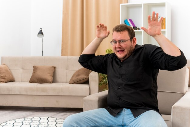 Bang volwassen Slavische man in optische bril zit op fauteuil met opgeheven handen in de woonkamer