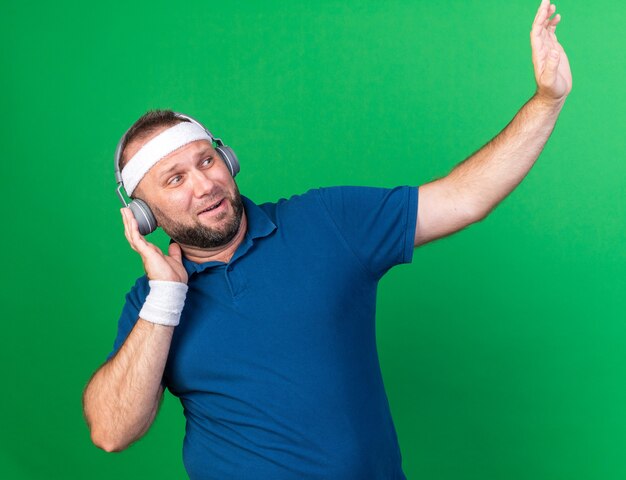 bang volwassen Slavische sportieve man op koptelefoon met hoofdband en polsbandjes kijkend naar de zijkant die hand omhoog steekt geïsoleerd op groene muur met kopieerruimte