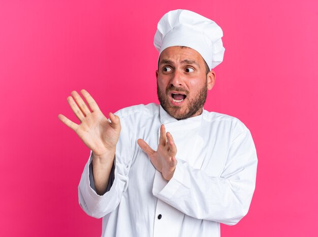 Bange jonge blanke mannelijke kok in uniform van de chef en pet die naar de zijkant kijkt en een weigeringsgebaar doet