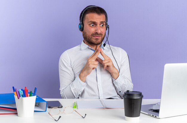Bange jonge mannelijke callcentermedewerker met een headset die aan tafel zit met kantoorhulpmiddelen die naar een laptop kijken