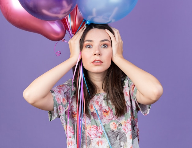 Bange jonge mooie vrouw met feestmuts met ballonnen greep het hoofd geïsoleerd op een blauwe muur
