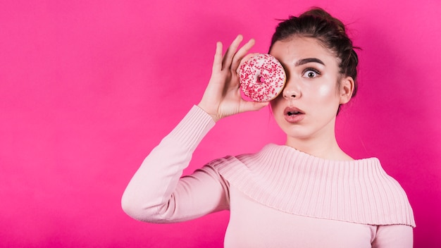 Bange jonge vrouw met donut over haar ogen tegen roze achtergrond