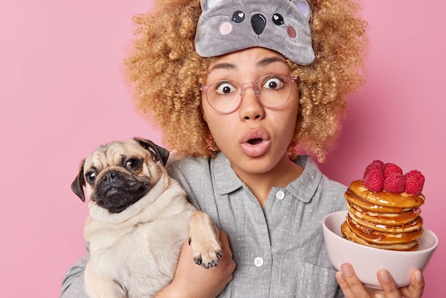 Bange jonge vrouw met krullend haar poseert met pannenkoeken met frambozen en mopshond die gaat ontbijten gekleed in pyjama en slaapmasker kan haar ogen geïsoleerd over roze achtergrond niet geloven