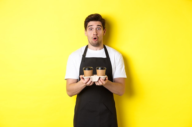 Barista serveert koffie, kijkt verbaasd naar de camera, draagt een zwarte schort, staande tegen een gele achtergrond.