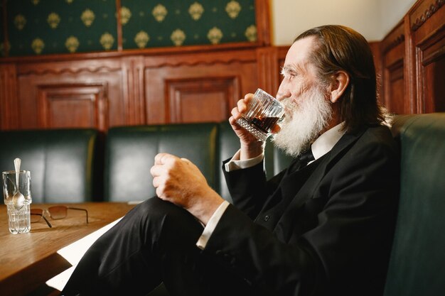 Bebaarde bejaarde zakenman. Man drinkt een whisky. Senior in zwart pak.