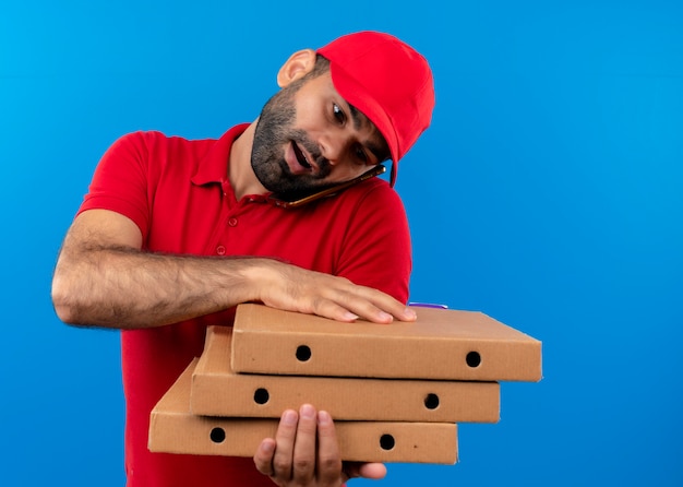 Bebaarde bezorger in rood uniform en pet bedrijf stapel pizzadozen praten op mobiele telefoon staande over blauwe muur