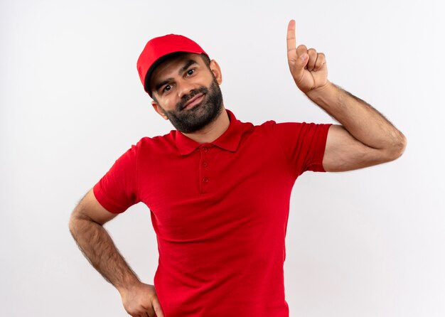 Bebaarde bezorger in rood uniform en pet omhoog met wijsvinger glimlachend vriendelijk staande over witte muur