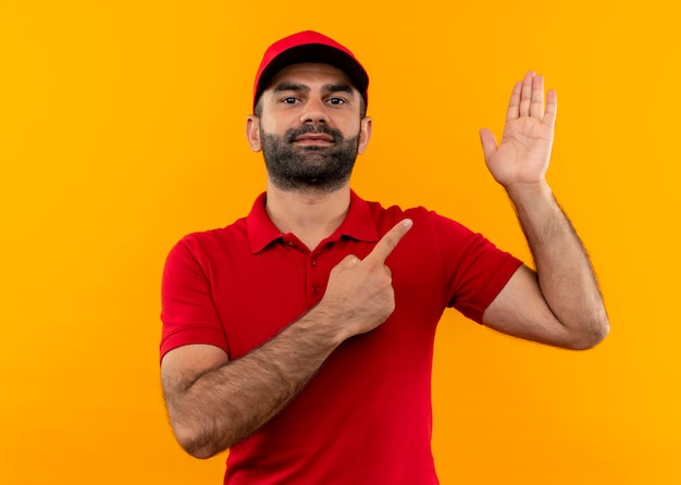 Bebaarde bezorger in rood uniform en pet wijzend met vinger naar arm staande over oranje muur