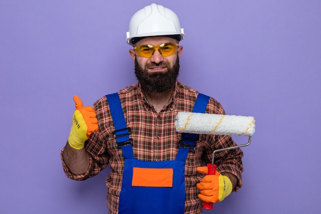 Bebaarde bouwer man in bouw uniform en veiligheidshelm dragen van rubberen handschoenen met verfroller kijken camera glimlachen tonen duimen omhoog permanent over paarse achtergrond