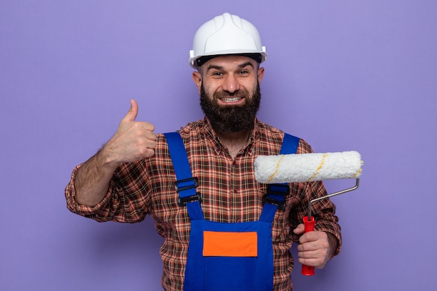 Bebaarde bouwer man in bouw uniform en veiligheidshelm met verfroller kijkend naar camera glimlachend vrolijk duimen opdagen staande over paarse achtergrond