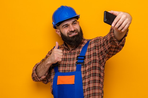 Bebaarde bouwman in bouwuniform en veiligheidshelm die selfie maakt met smartphone die vrolijk lacht met duimen omhoog