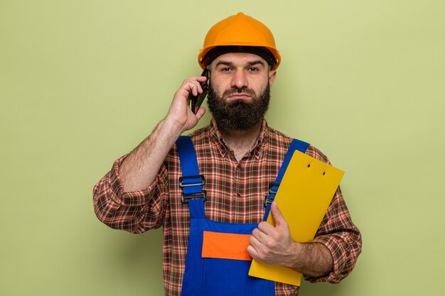 Bebaarde bouwman in bouwuniform en veiligheidshelm met klembord kijkend met een serieus gezicht pratend op mobiele telefoon