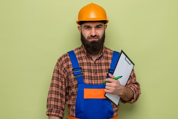 Bebaarde bouwman in bouwuniform en veiligheidshelm met klembord met pen die er zelfverzekerd uitziet