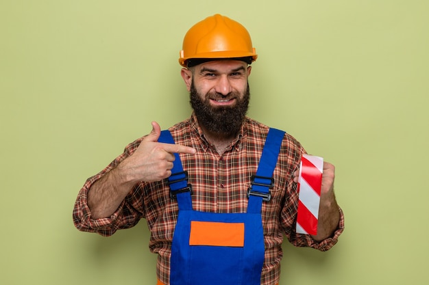 Gratis foto bebaarde bouwman in bouwuniform en veiligheidshelm met plakband die met de wijsvinger erop wijst en vrolijk lacht