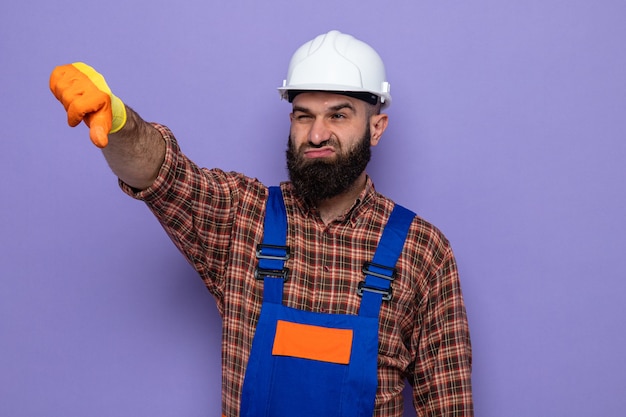 Bebaarde bouwman in bouwuniform en veiligheidshelm met rubberen handschoenen die opzij kijken en ontevreden zijn met duimen naar beneden