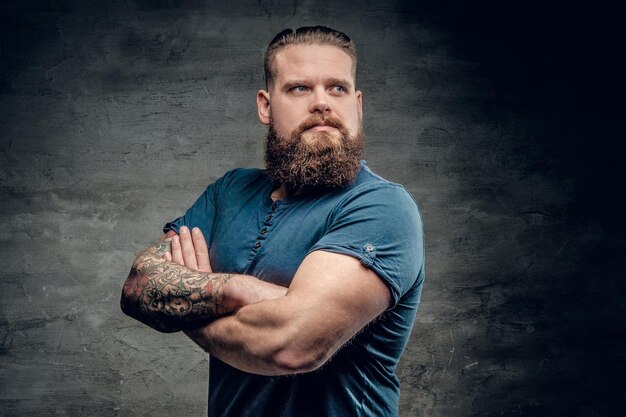 Bebaarde dikke man met tatoeage op armen poseren over grijze achtergrond.