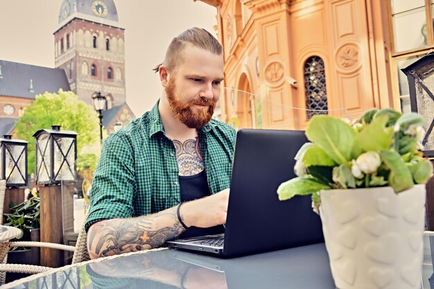 Bebaarde getatoeëerde man met laptop in een open straatcafé in de oude stad.