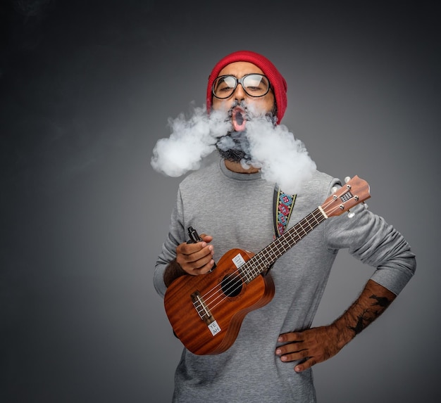 Bebaarde, informele man die een akoestische kleine gitaar vasthoudt en een sigaret rookt.