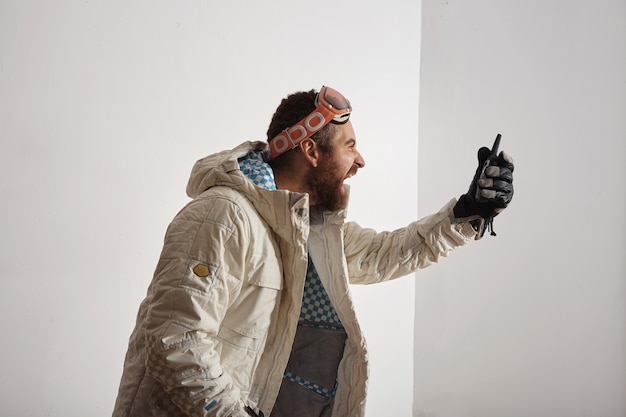 Bebaarde jonge man in snowboard jas en bril op zijn hoofd schreeuwen in walkie talkie voor hem, geïsoleerd op wit