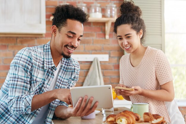 Bebaarde jongeman draagt geruit overhemd toont iets op tabletcomputer aan zijn vrouw die broodjes maakt