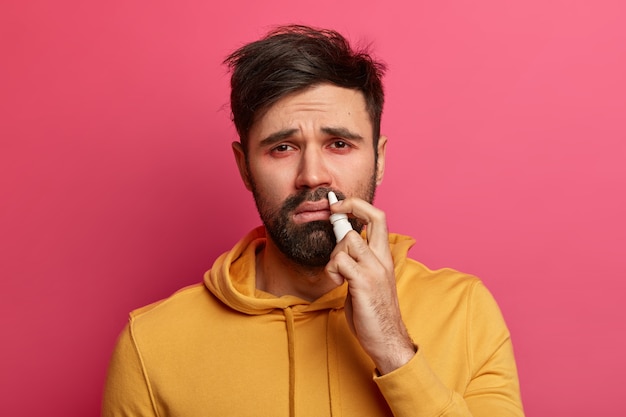 Bebaarde jongeman met rode ogen, loopneus en symptomen van griep of verkoudheid, spuit neus met druppels, geneest epidemie, gebruikt de beste remedie voor verstopte neus, draagt geel sweatshirt, probeert niet te niezen