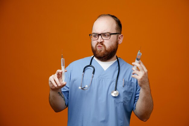 Bebaarde man arts in uniform met een stethoscoop om de nek met een bril die spuiten vasthoudt en er verward uitziet met twijfels over een oranje achtergrond