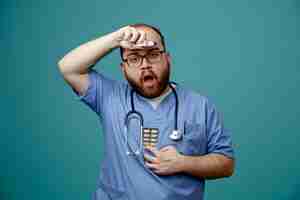 Gratis foto bebaarde man arts in uniform met stethoscoop om nek met bril met thermometer en pillen die verward en bezorgd over blauwe achtergrond staan