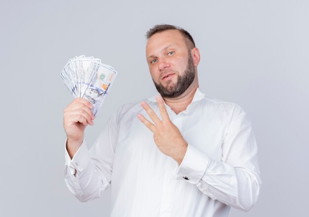 Bebaarde man die een wit overhemd draagt dat contant geld toont en met vingers omhoog wijst nummer drie glimlachend zelfverzekerd over witte muur