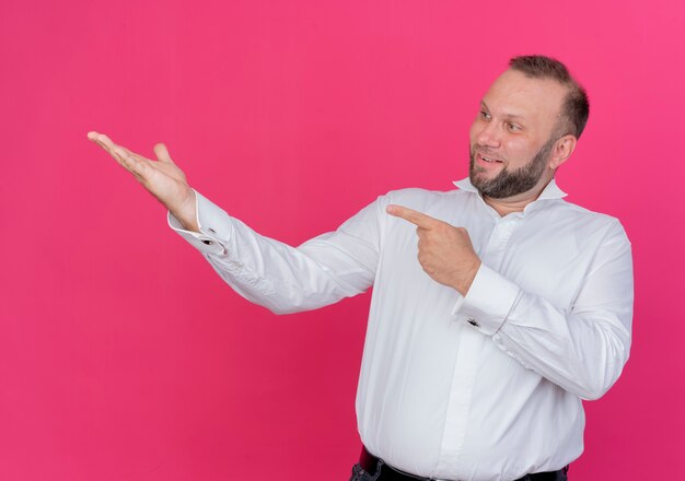 Bebaarde man die een wit overhemd draagt dat iets presenteert met een arm die met wijsvinger ernaar wijst glimlachend staande over roze muur