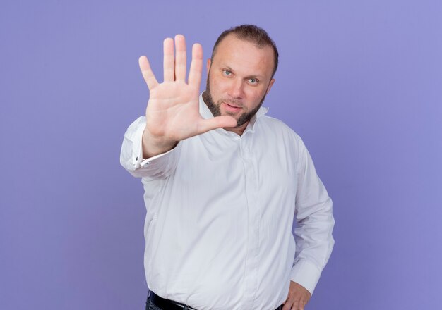 Bebaarde man die een wit overhemd draagt dat met ernstig gezicht kijkt en stopbord met hand maakt die zich over blauwe muur bevindt
