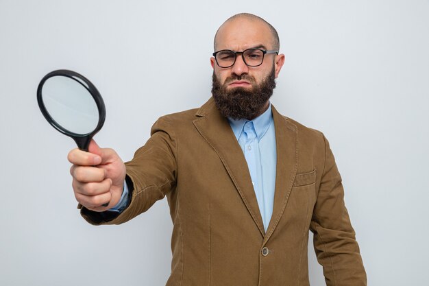 Bebaarde man in bruin pak met een bril die een vergrootglas vasthoudt en er doorheen kijkt met een serieus gezicht op een witte achtergrond