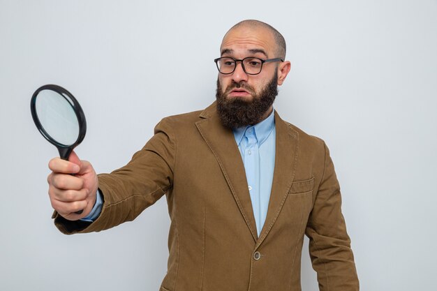 Bebaarde man in een bruin pak met een bril die een vergrootglas vasthoudt en er verward doorheen kijkt terwijl hij op een witte achtergrond staat