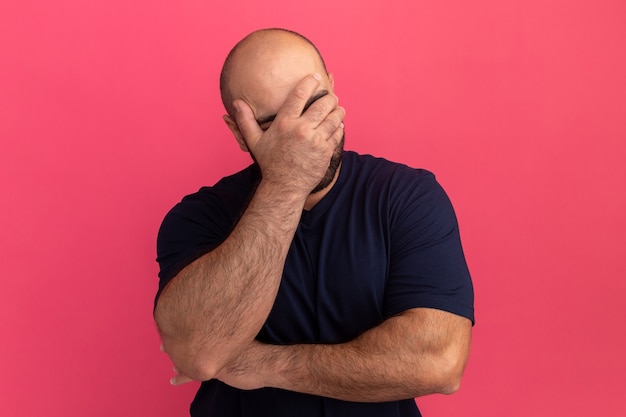 Bebaarde man in marine t-shirt die gezicht bedekt met hand op zoek verveeld en depressief staande over roze muur