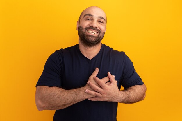 Bebaarde man in marine t-shirt lachend met handen op de borst dankbaar gevoel staande over oranje muur