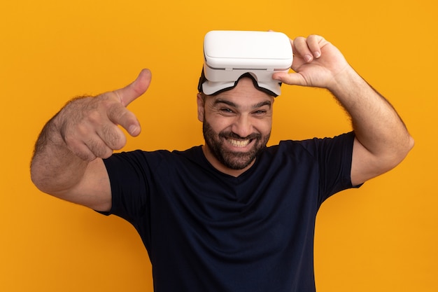 Bebaarde man in marine t-shirt met bril van virtual reality blij en opgewonden wijzend met wijsvinger staande over oranje muur