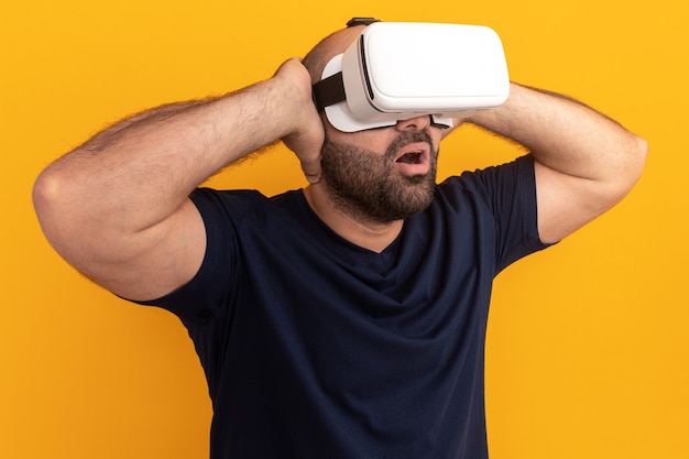 Bebaarde man in marine t-shirt met bril van virtual reality kijkt verbaasd en verrast staande over oranje muur