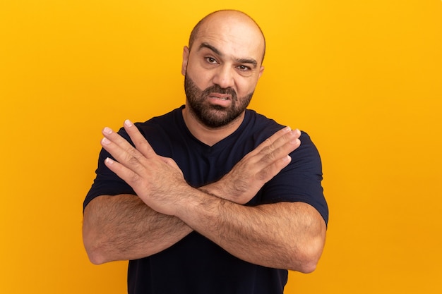 Bebaarde man in marine t-shirt met ernstig gezicht handen kruisen stop gebaar staande over oranje muur maken