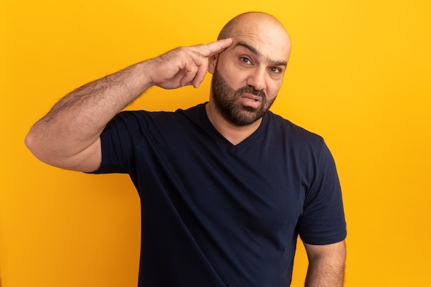 Bebaarde man in marine t-shirt met zelfverzekerde uitdrukking op slim gezicht wijzend met wijsvinger naar haar tempel staande over oranje muur