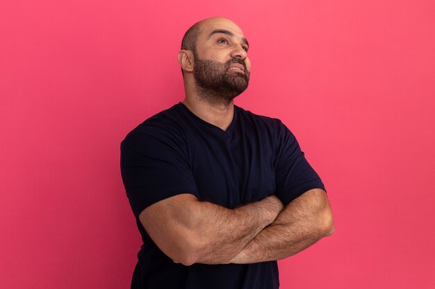 Bebaarde man in marine t-shirt opzoeken met ernstige peinzende uitdrukking met gekruiste armen staande over roze muur