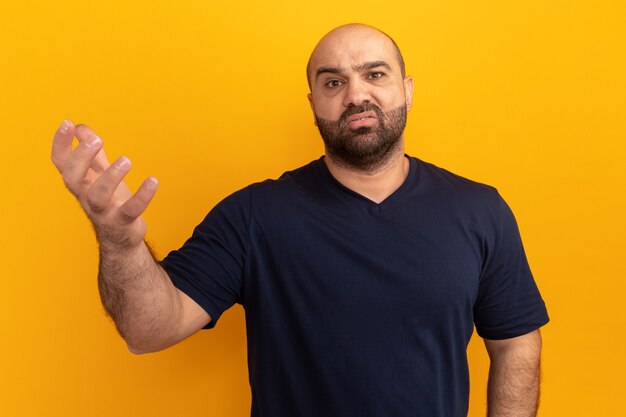 Bebaarde man in marineblauw t-shirt, verward en erg angstig, arm opheffend van ongenoegen en verontwaardiging staande over oranje muur