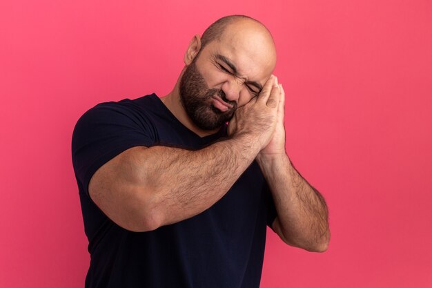 Bebaarde man in marineblauw t-shirt wil slapen slaapgebaar maken handpalmen tegen elkaar houden leunend hoofd op handpalmen staande over roze muur