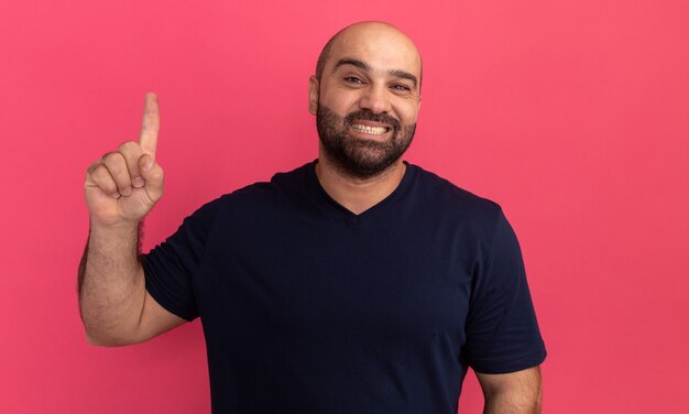 Bebaarde man in marinet-shirt met een grote glimlach op gezicht met wijsvinger die zich over roze muur bevindt