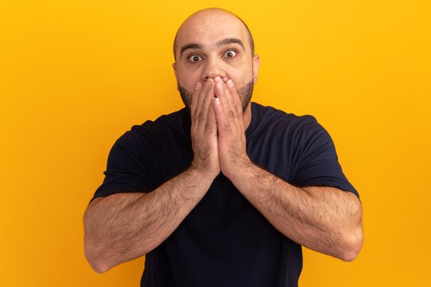 Bebaarde man in marinet-shirt verbaasd bedekkende mond met handen die zich over oranje muur bevinden