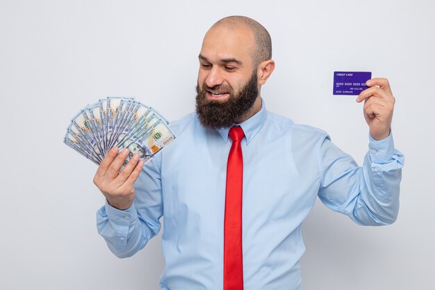 Bebaarde man in rode stropdas en blauw shirt met contant geld en creditcard kijkend naar geld gelukkig en tevreden glimlachend vrolijk staande over witte achtergrond