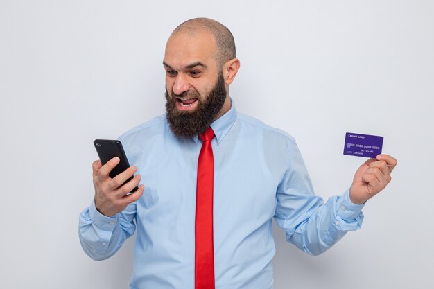 Bebaarde man in rode stropdas en blauw shirt met creditcard en smartphone die er blij en opgewonden naar kijkt en vrolijk lacht