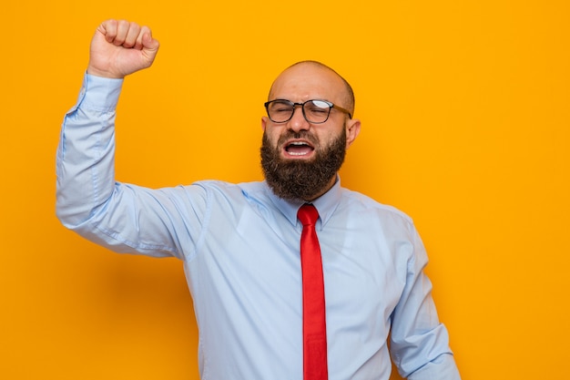 Bebaarde man in rode stropdas en blauw shirt met een bril, blij en opgewonden, gebalde vuist