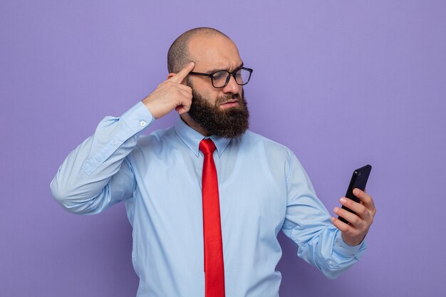 Bebaarde man in rode stropdas en blauw shirt met een bril die een smartphone vasthoudt en er verbaasd naar kijkt