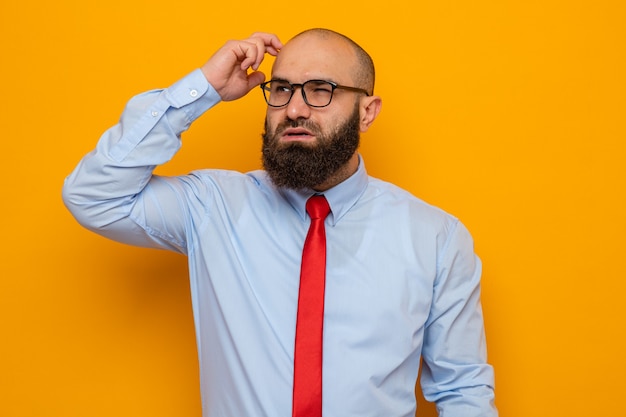 Bebaarde man in rode stropdas en shirt met bril opzij kijkend verbaasd op zijn hoofd krabben