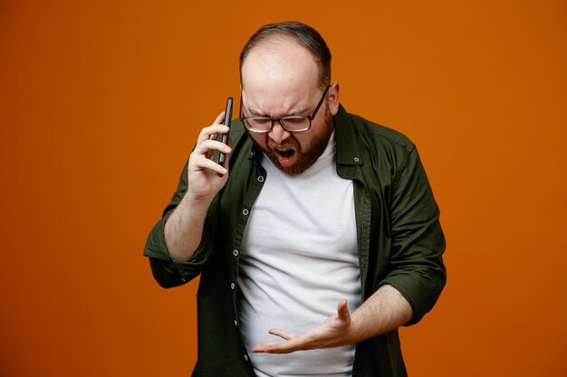 Bebaarde man in vrijetijdskleding met een bril die boos en gefrustreerd kijkt terwijl hij op een mobiele telefoon praat en schreeuwt over een oranje achtergrond